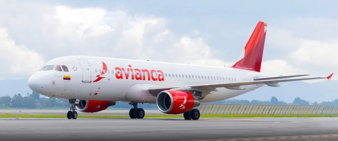 Aeronave Airbus A320 da Avianca taxiando na pista, com a fuselagem branca e cauda e motores em vermelho. A bandeira da Colômbia está pintada perto do nariz. O avião está sob um céu nublado, com colinas verdes ao fundo.