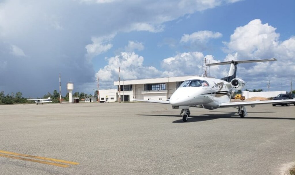 Jato executivo branco com detalhes em preto e dourado estacionado no pátio de um aeroporto. Ao fundo, vê-se um prédio administrativo com grandes janelas, uma caixa d'água branca e uma pequena aeronave monomotor distante na pista, sob um céu com nuvens carregadas.