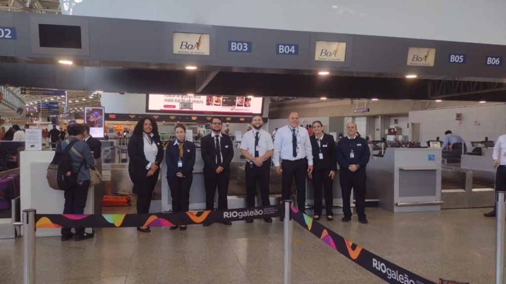 Três homens adultos sorriem lado a lado em um ambiente interno. Eles estão posicionados à frente de uma parede azul vibrante que exibe o logotipo branco da Universal Assistance.