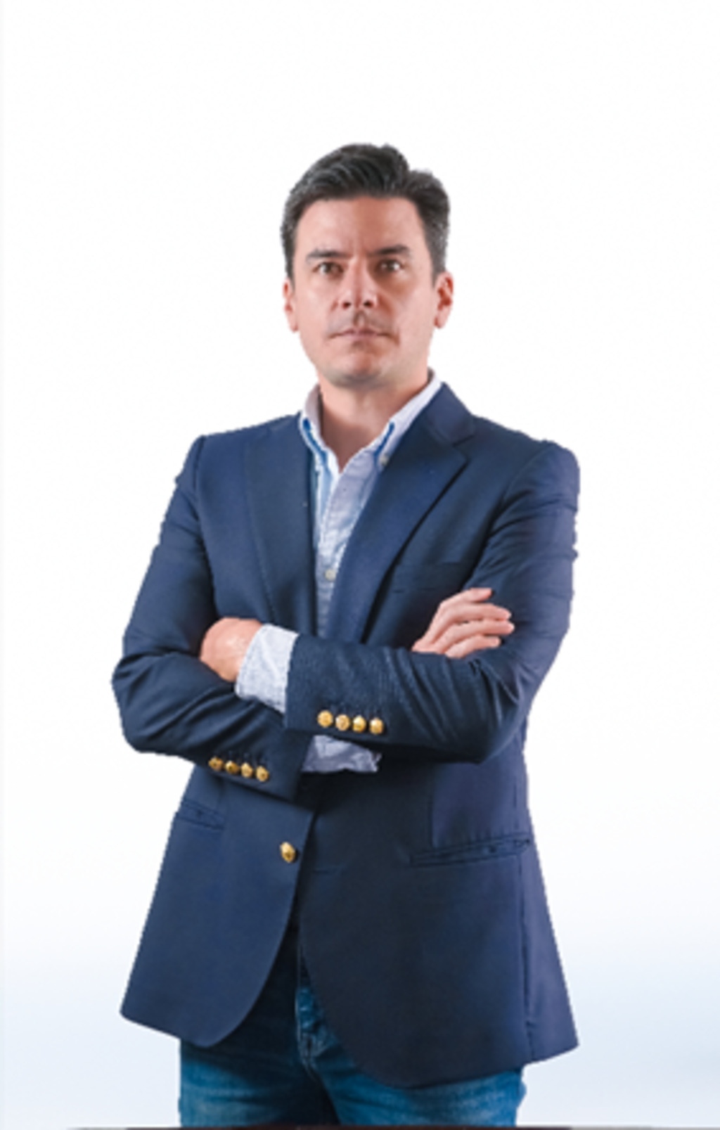 Retrato de um homem de cabelos escuros e expressão séria, posando com os braços cruzados contra um fundo branco. Ele veste um blazer azul-marinho com botões dourados, uma camisa social azul-clara e calça jeans.