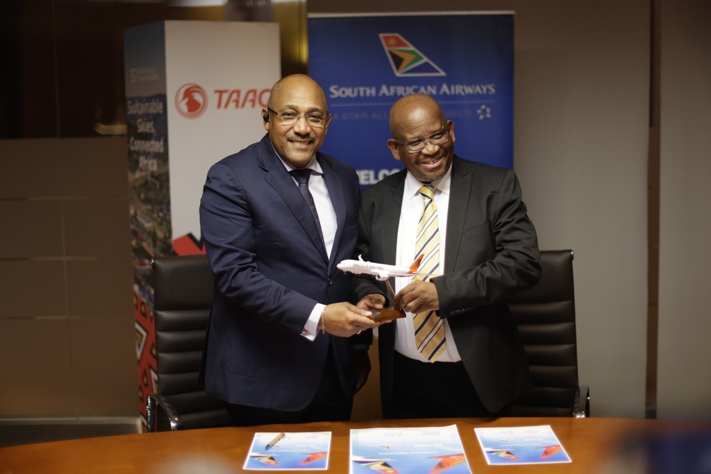 Duas pessoas em trajes de negócios trocam um modelo de avião; ao fundo estão logotipos da TAAG Angola Airlines e South African Airways