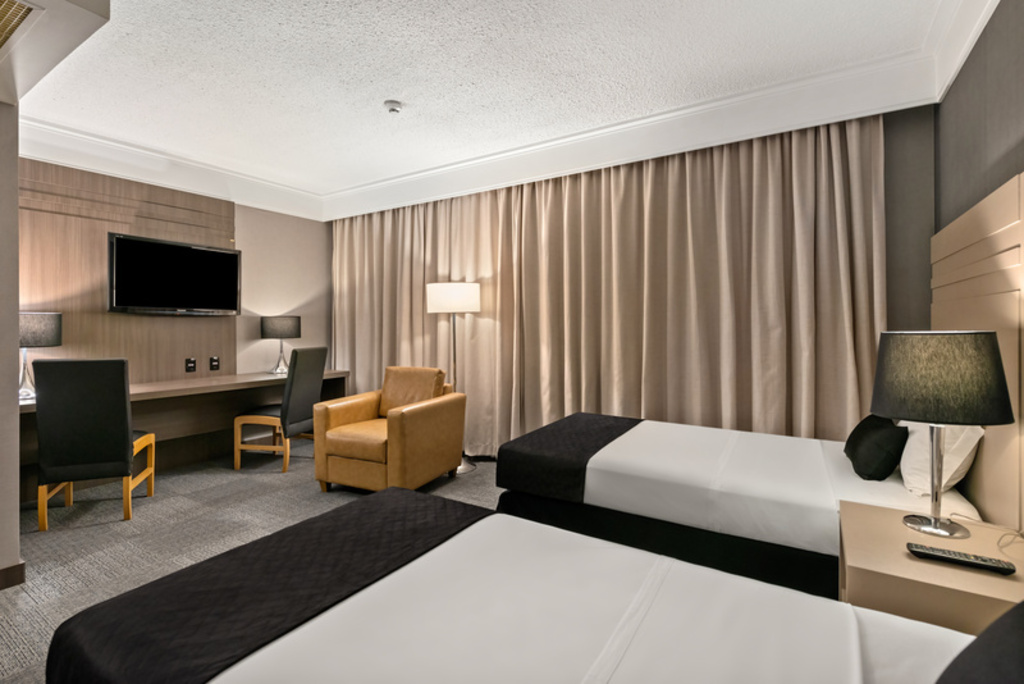 Quarto de hotel moderno com duas camas de solteiro dispostas em paralelo no primeiro plano, cobertas com lençóis brancos e mantas pretas nos pés. À direita, um criado-mudo bege sustenta um abajur de cúpula preta.