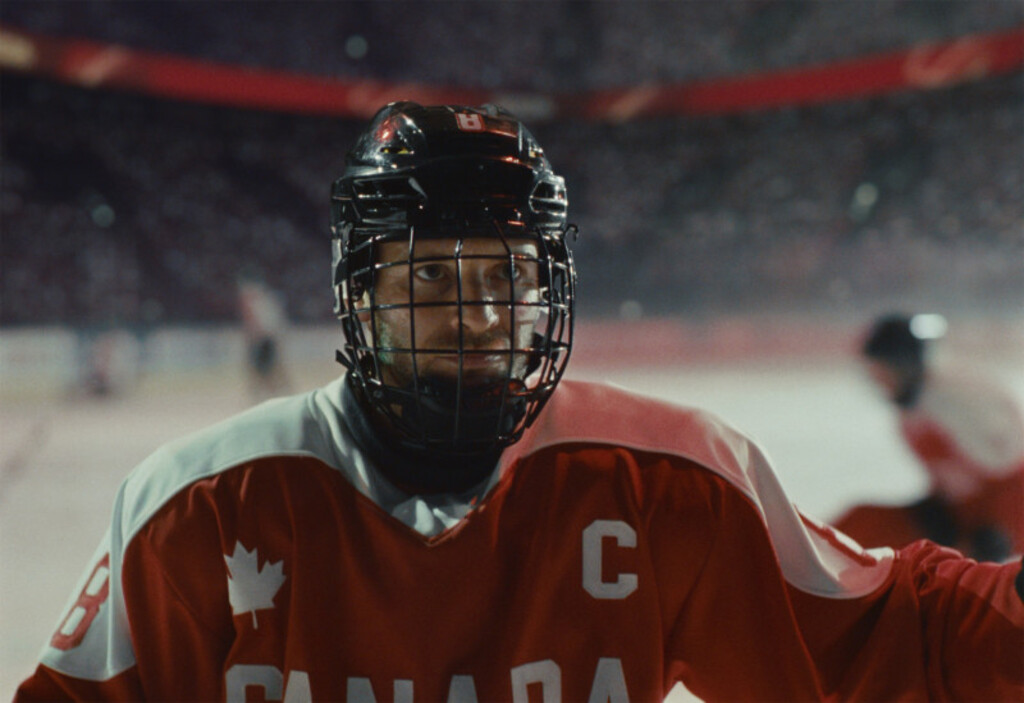 Retrato em close-up de um jogador de hóquei no gelo da seleção do Canadá, em destaque no centro da imagem. Ele está uniformizado e usa um capacete preto com grade de proteção facial, olhando fixamente para o lado.