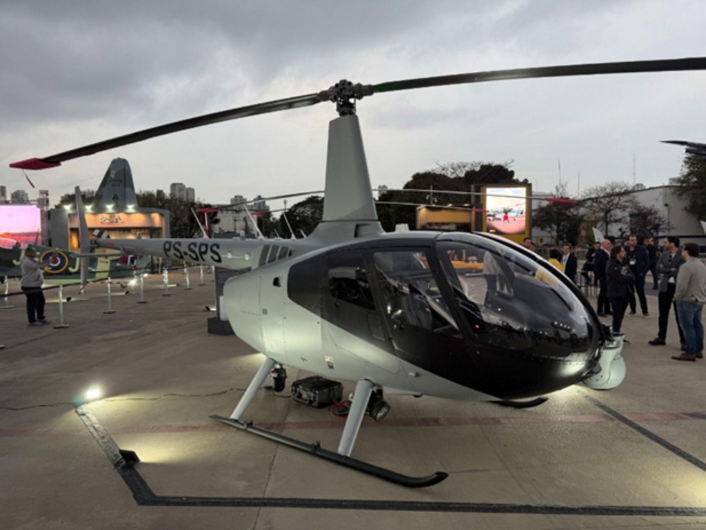Helicóptero modelo Robinson R66 de cor cinza, visto de perfil diagonal, estacionado em um pátio de eventos. A cabine é escura e a iluminação do chão destaca a parte inferior da aeronave. Céu nublado ao fundo com luzes de cidade e público visitante.
