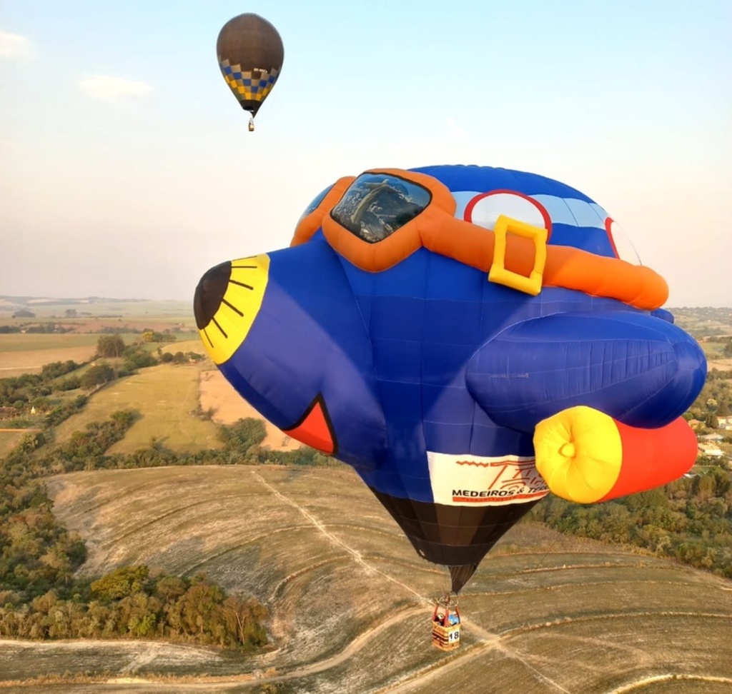 Fotografia aérea de um balão de ar quente com formato especial de avião azul cartoonizado sobrevoando uma paisagem rural. O balão possui detalhes em amarelo no bico, 