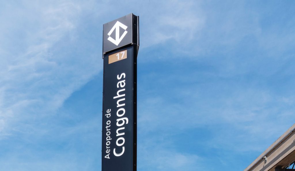 Fotografia em ângulo contra-picado de um totem de sinalização vertical preto sob um céu azul com nuvens leves. No topo do totem, há o logotipo do Metrô de São Paulo (um losango formado por duas setas brancas). Logo abaixo, um pequeno quadrado na cor ouro traz o número 