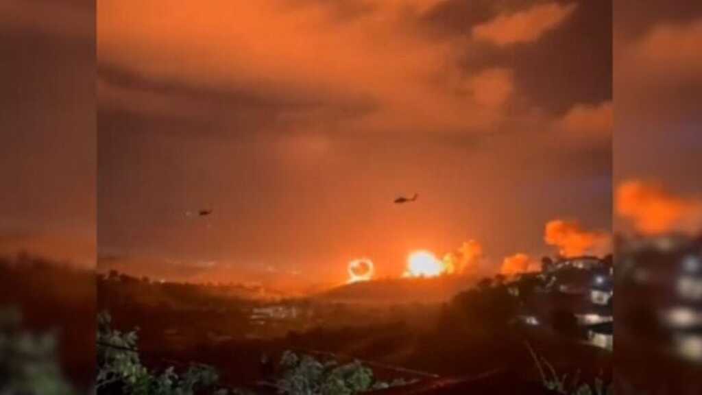 Fotografia noturna de uma paisagem montanhosa com explosões de fogo no horizonte. Dois helicópteros em silhueta sobrevoam a área sob um céu escuro e carregado. No primeiro plano, luzes de uma área urbana e vegetação aparecem na penumbra.