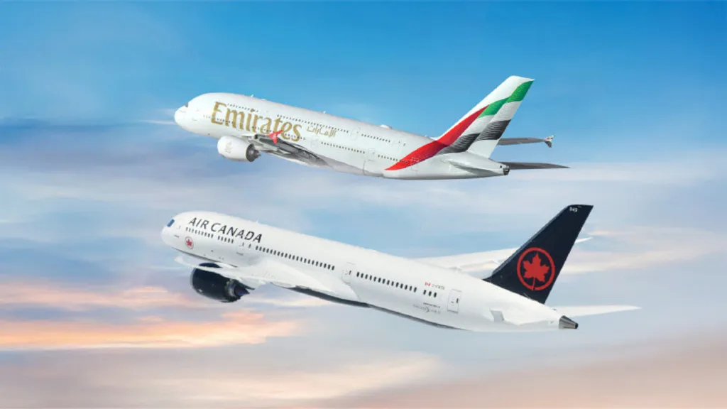 Uma imagem composta mostrando duas grandes aeronaves comerciais voando juntas no céu, uma acima da outra. Na parte superior, está um Airbus A380 da Emirates (com dois andares de janelas), branco com o logotipo dourado e a cauda com as cores da bandeira dos Emirados Árabes Unidos. Na parte inferior e um pouco à frente, voa um Boeing 787 Dreamliner da Air Canada, com fuselagem branca e uma cauda preta marcante contendo a folha de bordo vermelha (logotipo da companhia). O fundo é um céu azul suave com tons de pôr do sol no canto inferior.