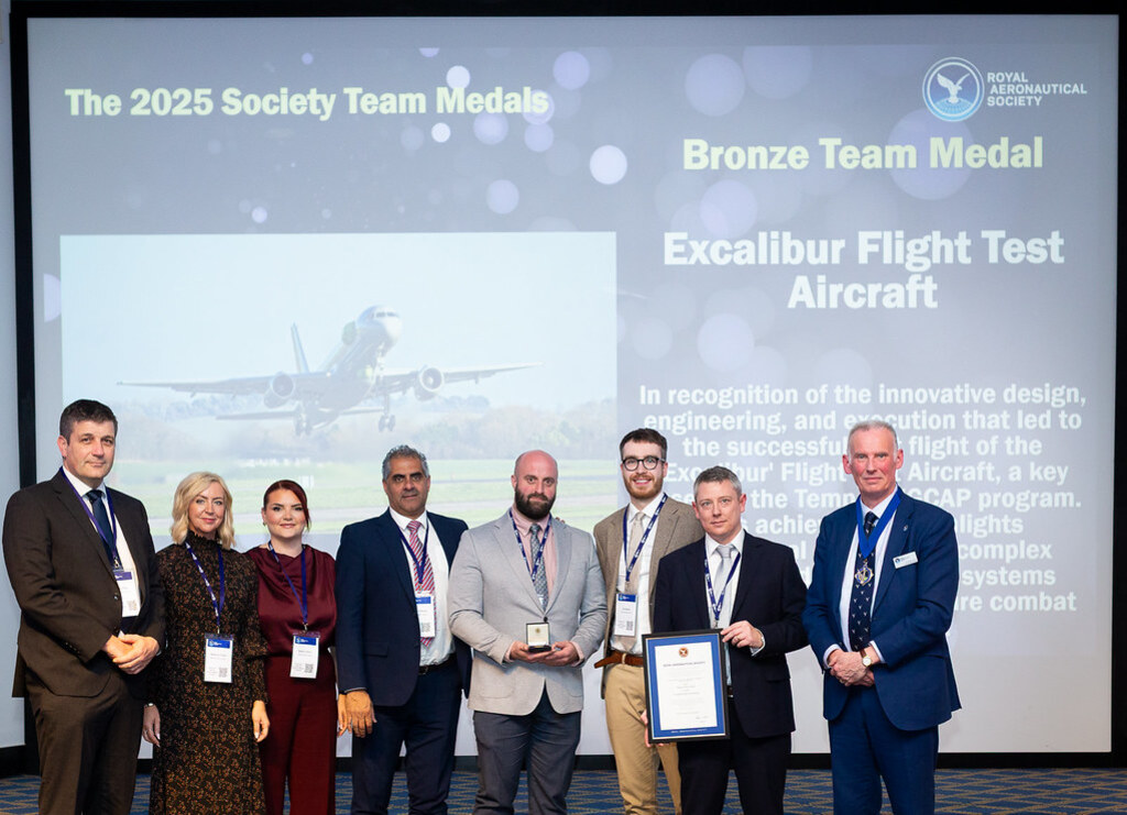 Uma foto em um palco de premiação mostra oito pessoas em trajes formais. Eles estão posando em frente a uma grande tela de projeção que exibe o texto 