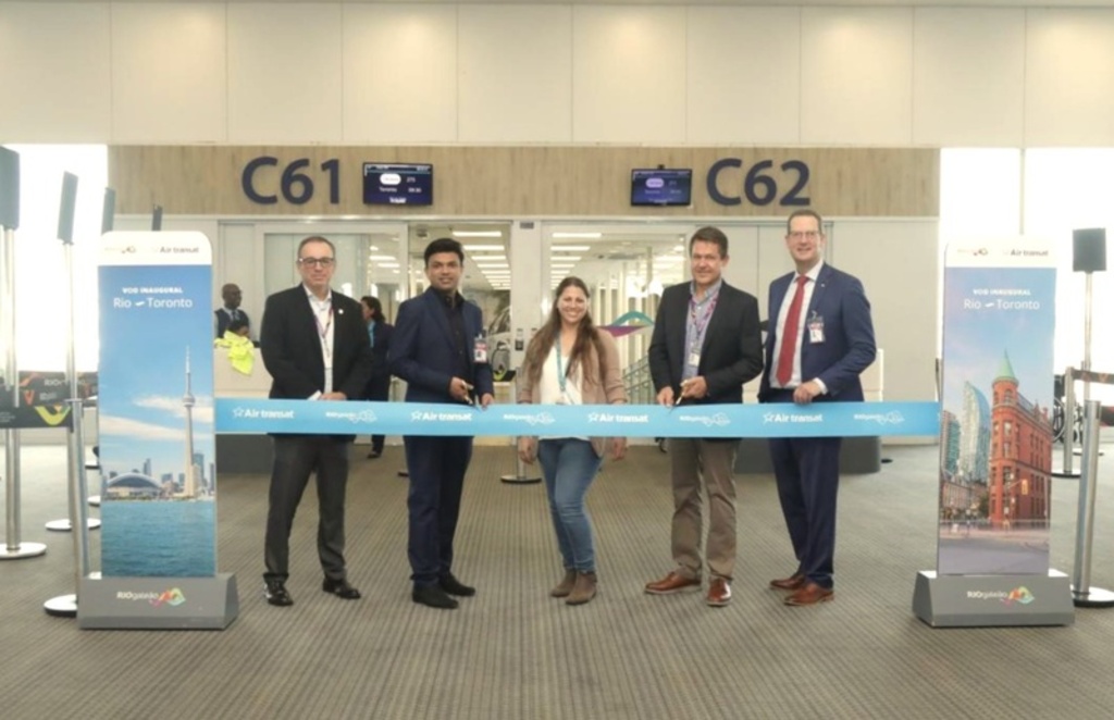 Cinco pessoas (quatro homens de terno e uma mulher de calça jeans e blazer) estão alinhadas atrás de uma longa fita azul claro com o logotipo da Air Transat. Dois dos homens seguram tesouras, prontos para cortar a fita.
