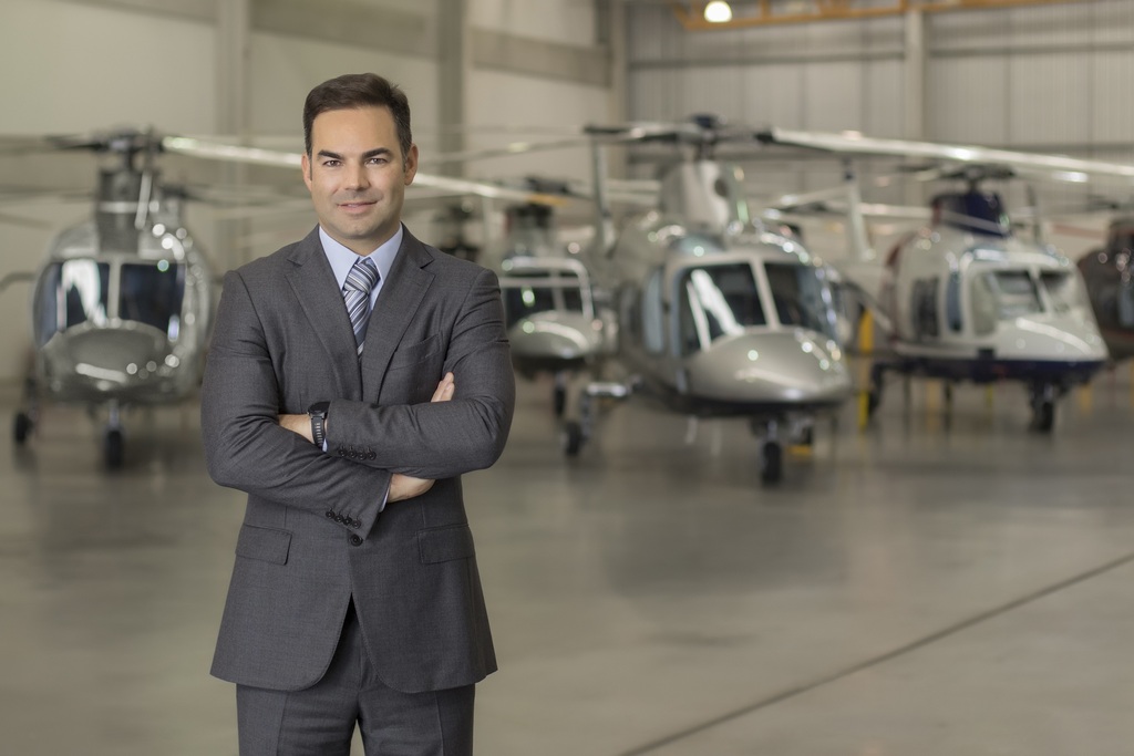 Homem de pele clara e cabelos castanhos curtos, vestindo um terno cinza escuro, camisa azul clara e gravata listrada, posa de braços cruzados e sorrindo discretamente para a câmera. Ele está posicionado em primeiro plano, no centro de um hangar de aeronaves. Ao fundo, de forma levemente desfocada, aparecem pelo menos três helicópteros executivos modernos estacionados sobre o piso de concreto polido do hangar.