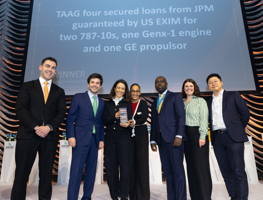 Um grupo de sete profissionais (quatro homens e três mulheres) posa sorridente em um palco durante uma premiação. Uma das mulheres ao centro segura um troféu com a inscrição 2026 Awards. Ao fundo, um telão exibe o texto sobre empréstimos da TAAG garantidos pelo US EXIM. Os homens vestem ternos e as mulheres usam trajes sociais formais. Nas laterais, há totens brancos com o logotipo da Airline Economics.