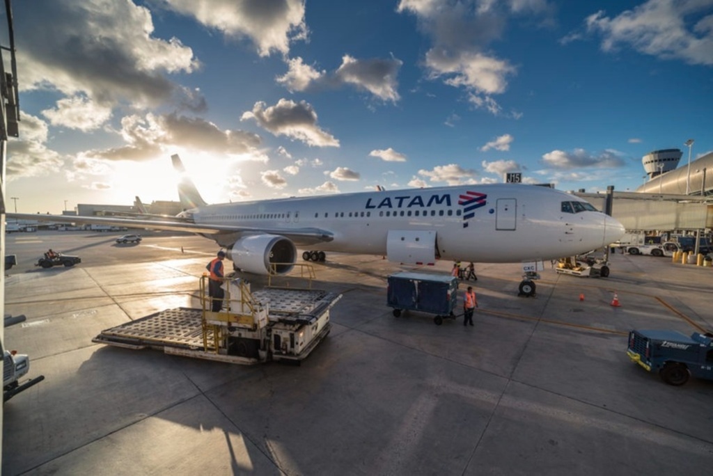 Avião da LATAM estacionado no pátio de um aeroporto durante o entardecer. A aeronave está cercada por equipamentos de suporte terrestre e funcionários em operação de carga. O sol brilha ao fundo entre nuvens esparsas, iluminando a pista.