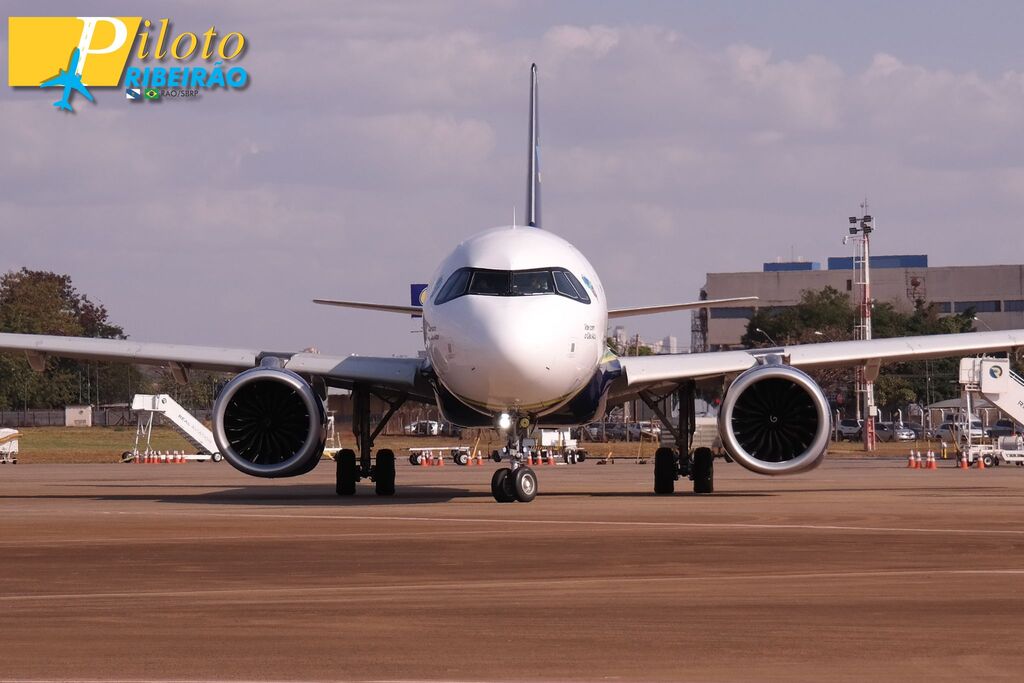 Vista frontal de uma aeronave estacionada na pista. O foco está no nariz do avião, cockpit e nas duas grandes turbinas sob as asas. Ao fundo, estruturas do aeroporto e escadas de embarque sob um céu claro.