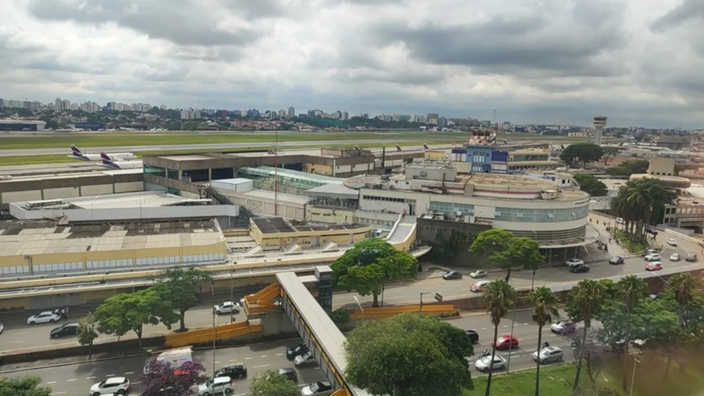 Vista aérea do Aeroporto de Congonhas em São Paulo sob um céu nublado. A imagem mostra o terminal de passageiros com sua arquitetura arredondada característica e a torre de controle ao fundo. No primeiro plano, uma avenida movimentada com carros e uma passarela amarela para pedestres. Ao fundo, a pista do aeroporto com aviões comerciais estacionados e o horizonte da cidade com diversos edifícios.