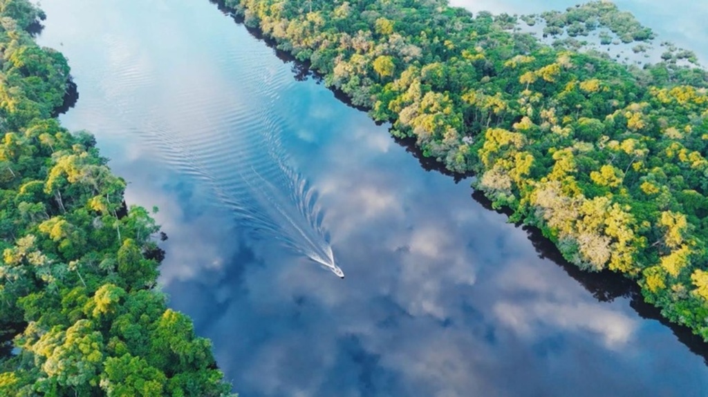 Rio da Amazônia ou similar visto de cima, com densa floresta verde nas margens. Um barco branco atravessa a água, que reflete o céu com nuvens