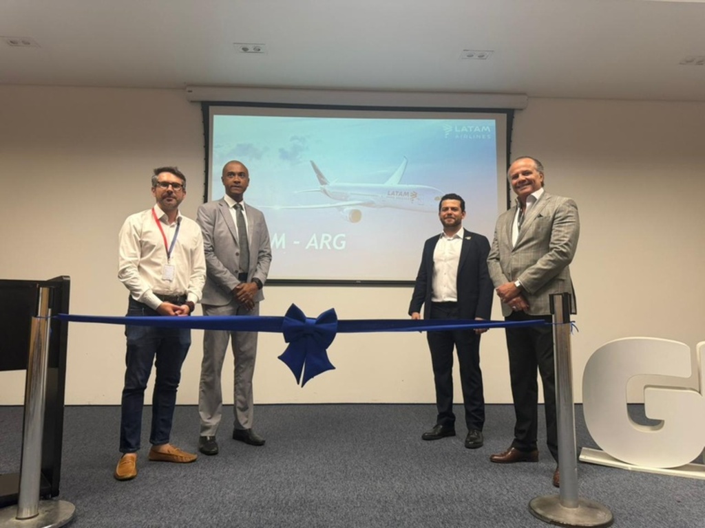 Quatro homens posam atrás de uma fita inaugural azul com um laço; ao fundo, um telão exibe uma imagem de uma aeronave da LATAM Airlines. No canto inferior direito, parte de uma grande letra 