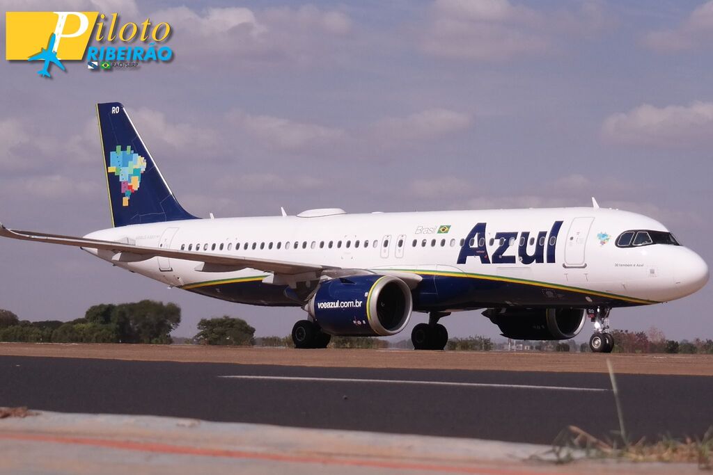 Vista lateral de um avião da Azul Linhas Aéreas taxiando em uma pista asfáltica sob a luz do dia. A aeronave é predominantemente branca com a parte inferior da fuselagem em azul-marinho, separada por faixas delgadas em verde e amarelo.
