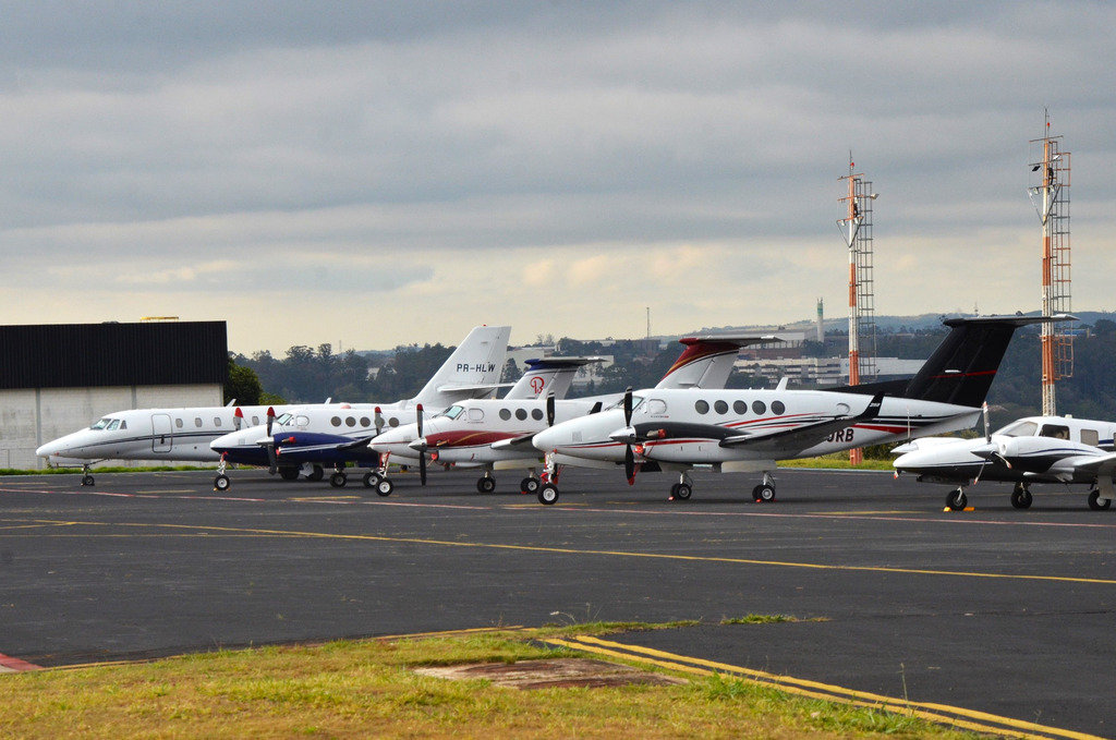 Várias aeronaves executivas de pequeno e médio porte estacionadas em um pátio de aeroporto sob um céu nublado, com torres de sinalização ao fundo.