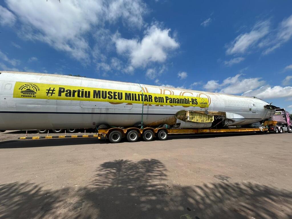 A fuselagem de um grande avião de passageiros (provavelmente um Boeing 727 ou similar), sem as asas e a cauda, está sendo transportada em uma carreta tipo prancha baixa de múltiplos eixos. A fuselagem é branca e tem uma faixa amarela horizontal com a inscrição em letras escuras: '# Partiu MUSEU MILITAR em Panambi-RS'. A seção central da aeronave está com um painel aberto, revelando a estrutura interna. Um caminhão de transporte de cor rosa e preto pode ser visto na frente. O transporte ocorre sob um céu azul claro com nuvens brancas, em uma área com solo de terra e cascalho.
