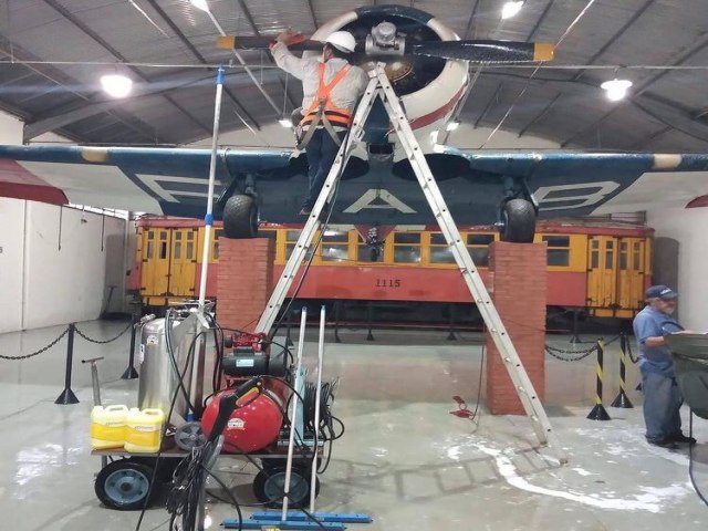 Dois homens realizando a manutenção ou limpeza da hélice e do motor radial de um avião histórico bimotor (provavelmente um Consolidated PBY Catalina) dentro de um hangar ou museu. Um dos homens está em cima de uma escada de alumínio, usando capacete e cinto de segurança. Abaixo da aeronave, há um antigo bonde ou carro de trem amarelo e vermelho em exibição. No piso, há um carrinho com equipamento de limpeza (mangueiras, reservatório e galões amarelos de produto) e outro homem é visível à direita, de costas.