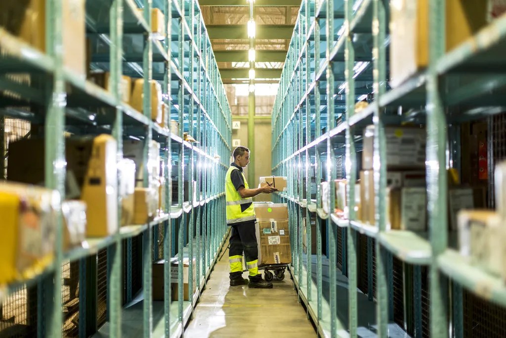 Um funcionário de armazém, vestindo um colete de segurança verde-limão e calças escuras, está em pé em um corredor estreito entre altas estantes metálicas repletas de caixas. Ele está manuseando uma pequena caixa de papelão, que parece estar colocando ou tirando de uma pilha em um carrinho de transporte no chão. A perspectiva é do fundo do corredor, com as prateleiras se estendendo para o primeiro plano, criando um túnel de perspectiva e focando no trabalhador. A luz natural entra pela abertura no final do armazém.