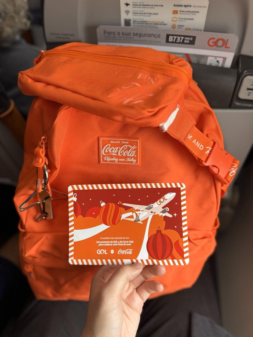 Uma mão segura um cartão festivo em frente a uma mochila laranja vibrante da Coca-Cola, dentro da cabine de um avião da GOL. O cartão apresenta uma ilustração de um avião da GOL voando entre enfeites de Natal e o texto: 